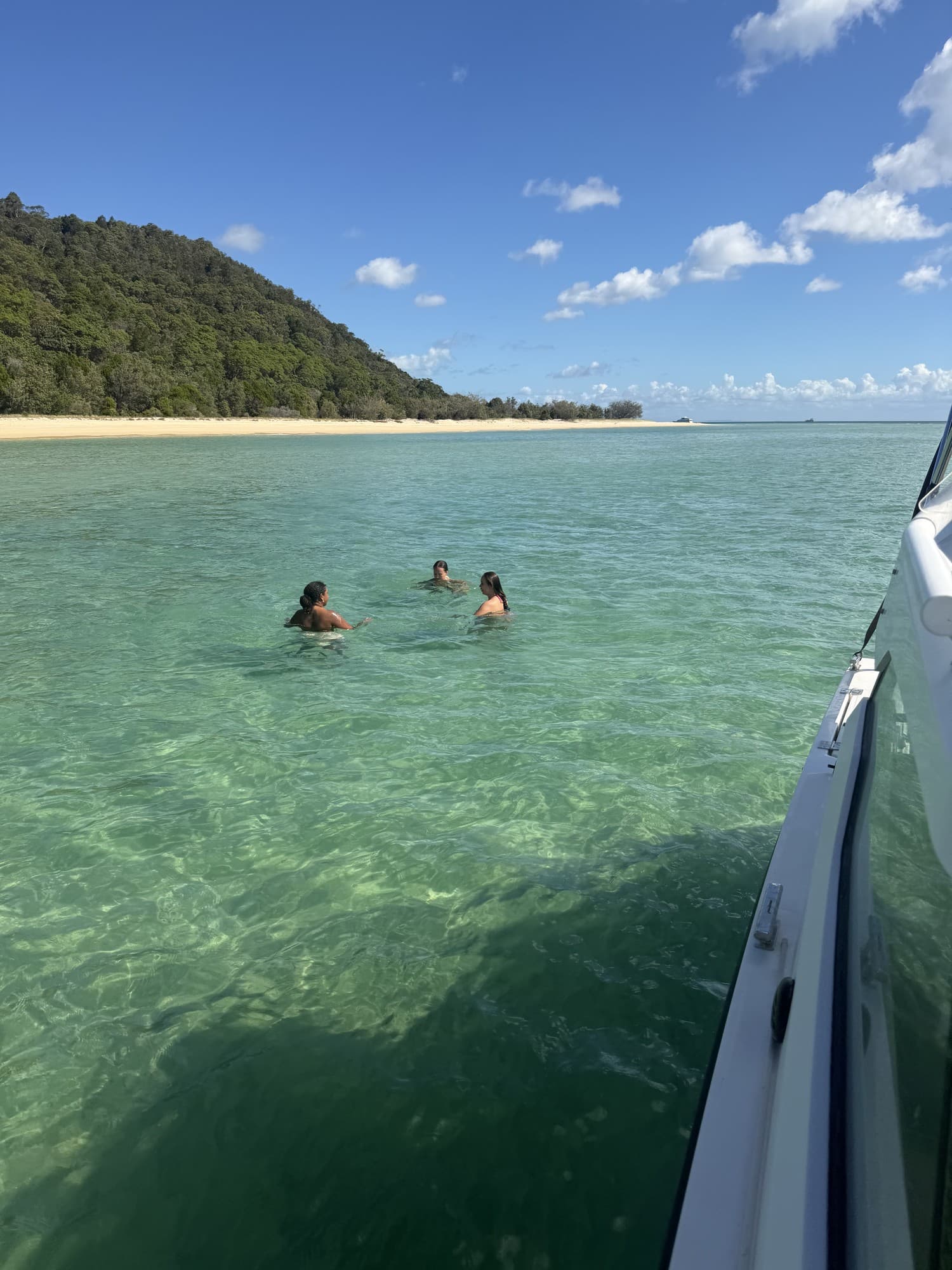 Two guests swimming in clear shallow water beside the boat, a wooded island shore behind